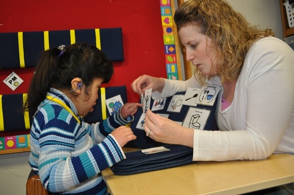A young girl points to a picture symbol that her teacher is holding