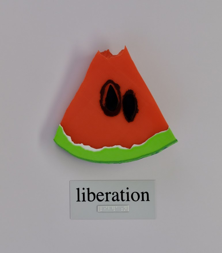 A tactile art piece of a watermelon slice with two big seeds and the words "liberation" underneath it in both print and braille