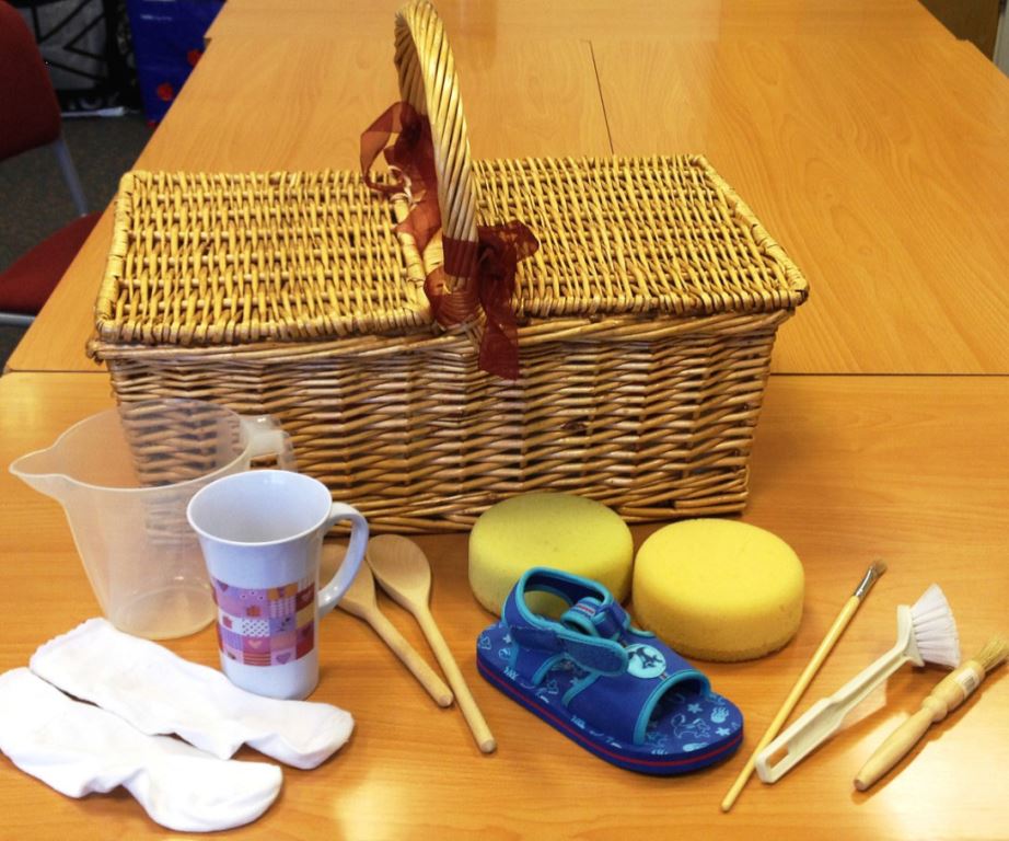 Basket of everyday objects, including socks, shoe, mug, measuring cup, wooden spoons, sponges, paintbrush