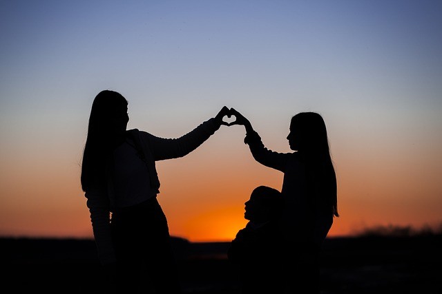 Two girls in shadow sitting at sunset holding hands into a heart shape.